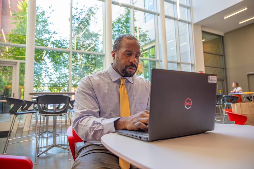 Male wearing a shirt and tie, seated at a table in front of a floor-to-ceiling window, is working on a laptop.