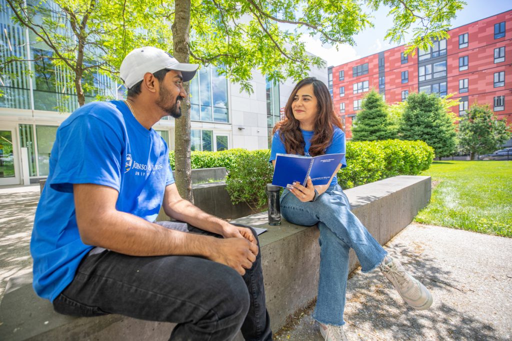 A male and a female, both wearing blue Johnson & Wales t-shirts, sit together on a low wall outdoors in a campus courtyard.