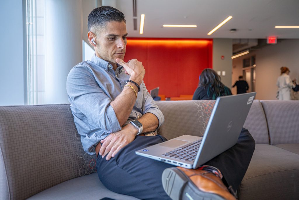 Young man seated in a common area in JWU's Bowen Center in Providence, looking thoughtful as he views his laptop screen.