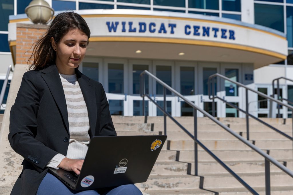 Young woman seated on the steps in front of the Wildcat Center on JWU's Harborside campus, viewing her laptop screen.