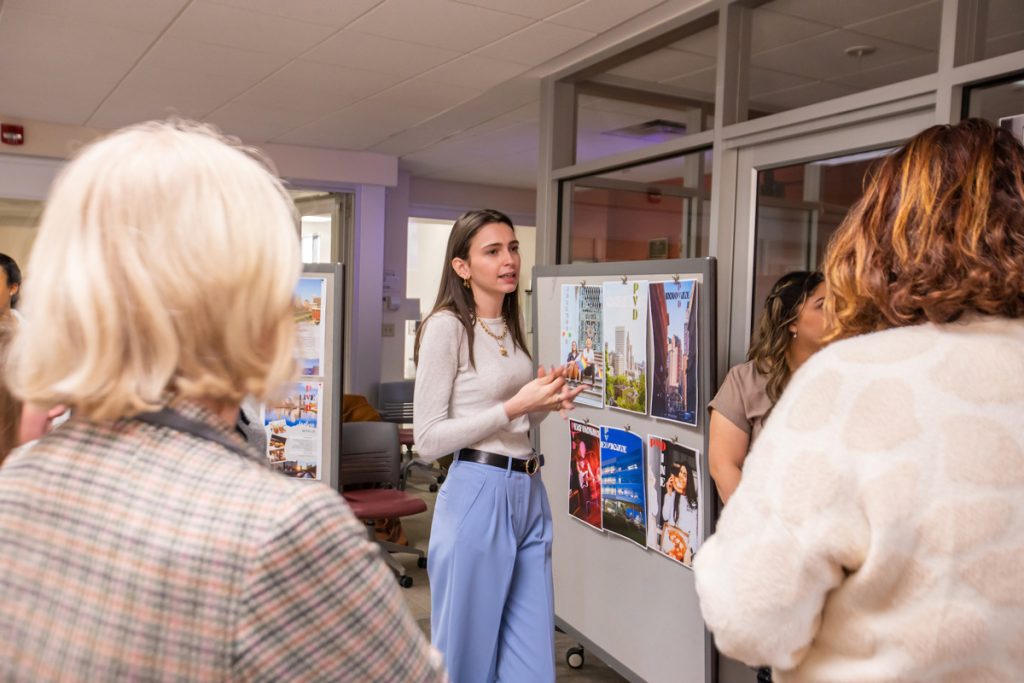 Adult female students grouped around one adult female student, who is presenting her work in the marketing lab.