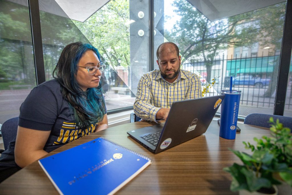 Female student and male advisor seated at a table, behind which is a window looking out to the street, view a laptop screen together.