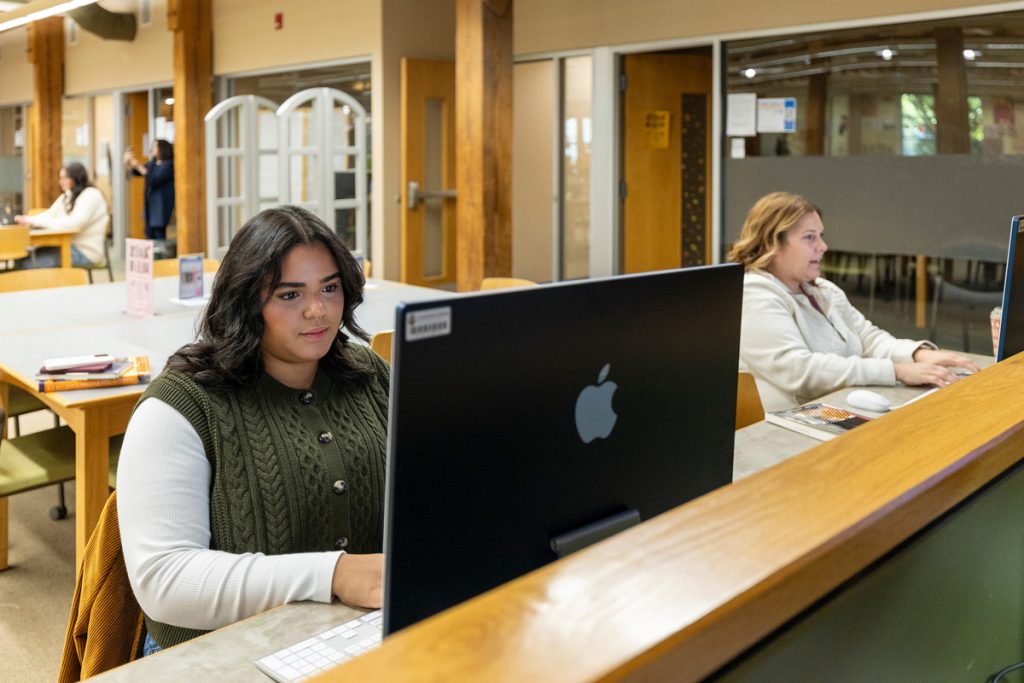 Two students in a university library are each looking at their laptop screens.