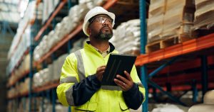 A warehouse worker in a yellow high-visibility jacket, white hard hat, and safety glasses holds a tablet while looking up at tall storage racks stocked with palletized goods