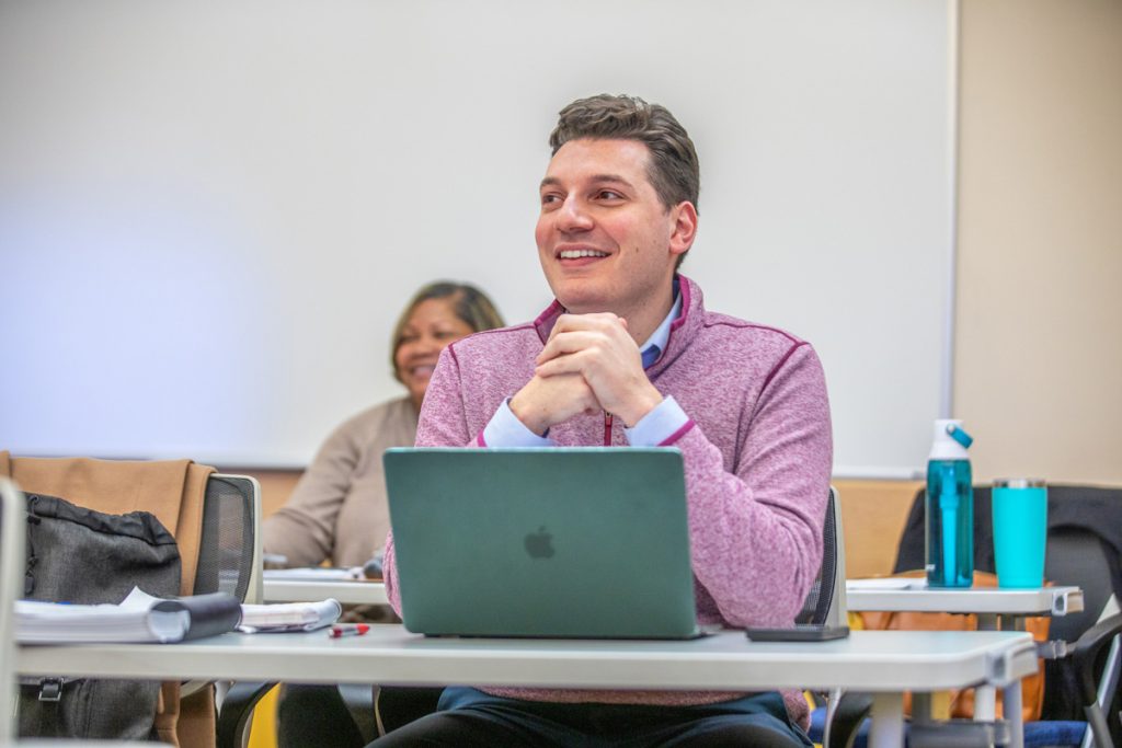 A male adult student seated at a table in a classroom has his hands clasped above his laptop screen and is smiling as he looks toward the front of the room.