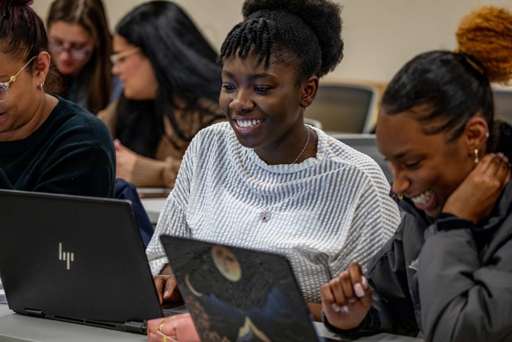 Two women of color in a classroom with other adult students are smiling as they view their laptop screens together.