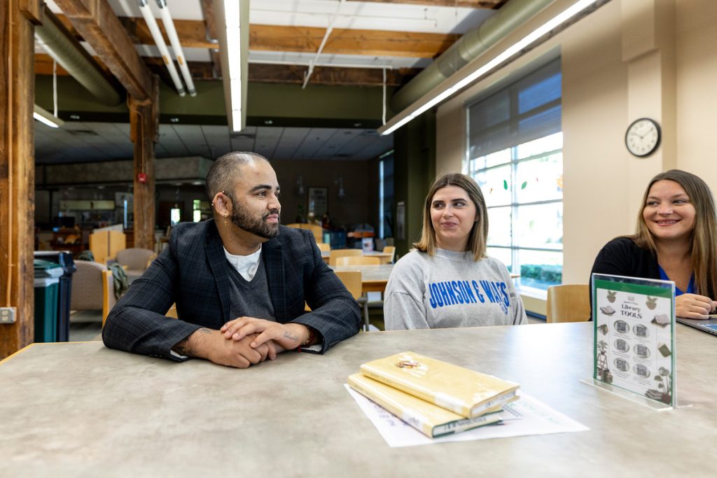 One male and two female adult students seated at a table in a library are smiling at one another as they talk together.