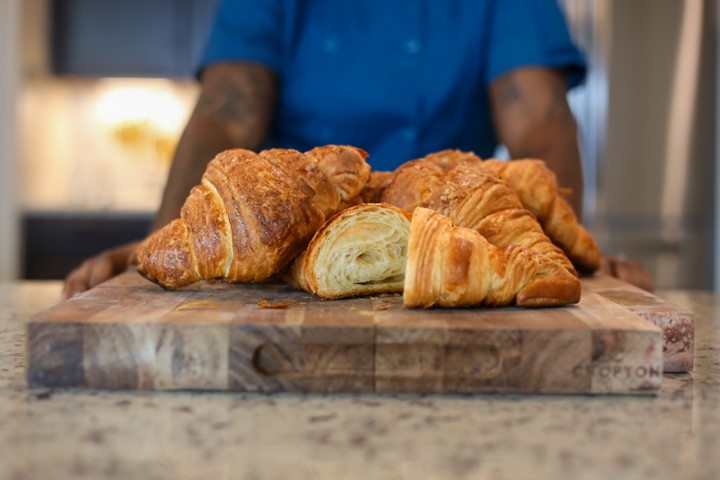 Freshly baked croissants on a wooden board behind which Direka Gordon's torso and arms are visible in soft focus.