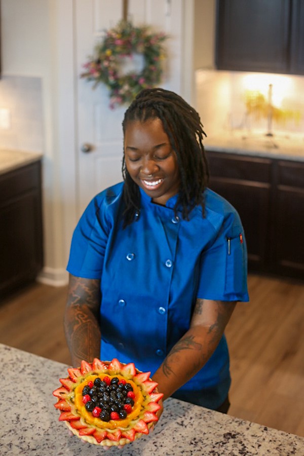 Direka Gordon wearing her blue chef's shirt, standing in her kitchen and smiling as she looks down on a freshly baked fruit tart she is holding.