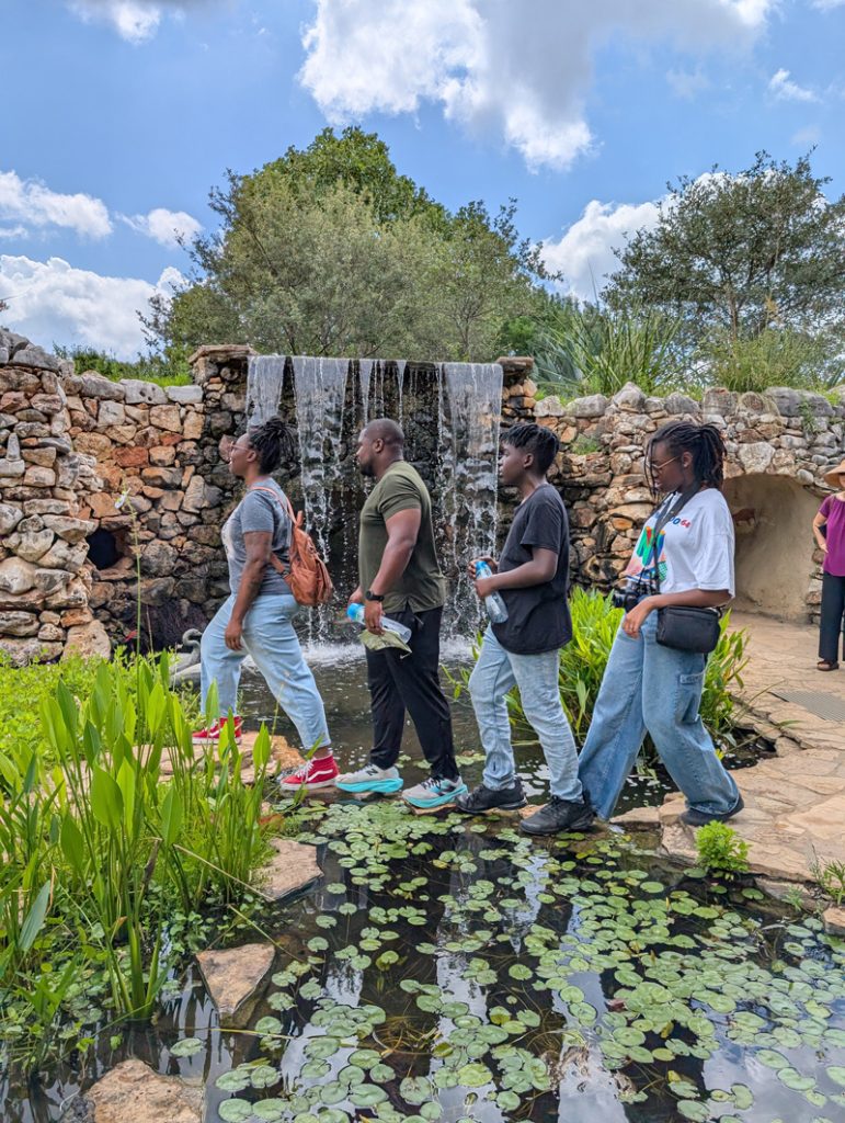 Direka Gordon and her family walking in single file on stepping stones across a stream from a waterfall, which is seen in the background, in imitation of the Beatle's Abbey Road album cover.