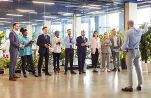 A multicultural group of hospitality professionals stands in a modern, plant-filled office space listening to leadership