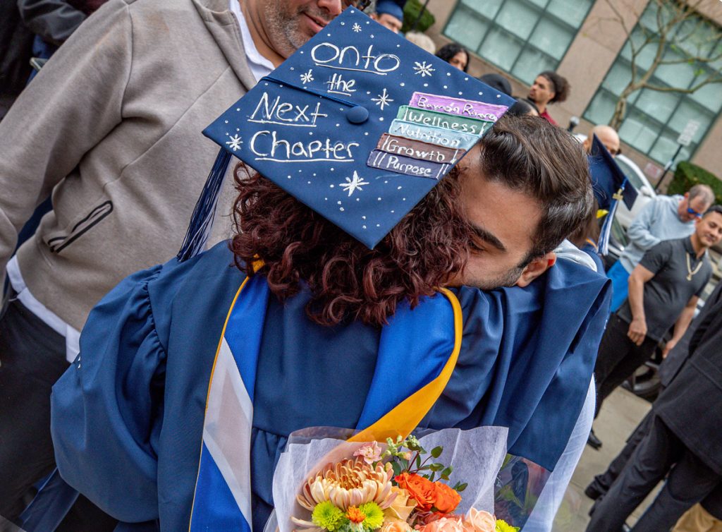 A view of the back of a young female graduate, wearing a cap decorated with the words, Onto the Next Chapter," and a young male wearing street clothes and holding flowers embrace.