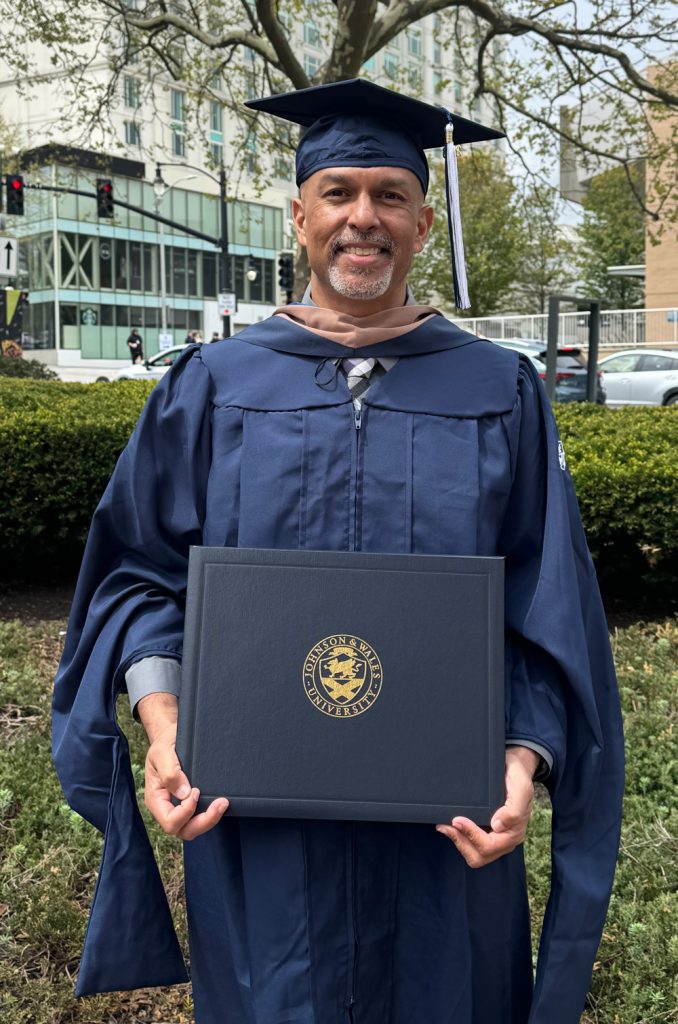 Ajay Patel. MD, outdoors in cap and gown and holding his diploma, after the commencement ceremony at the Amica Mutual Pavilion.