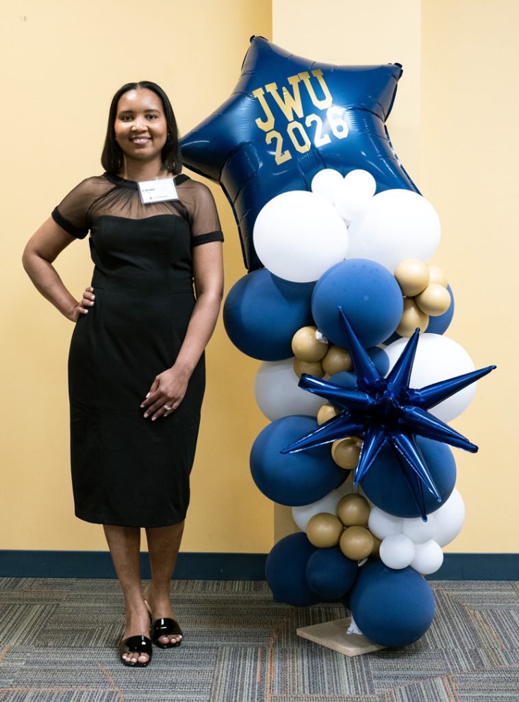 Djelisa Daveiga posing next to the blue and white balloon decoration embossed with JWU 2026 in gold letters, at the JWU Online 2026 Senior Toast held in the Wildcat Center on the afternoon before commencement.
