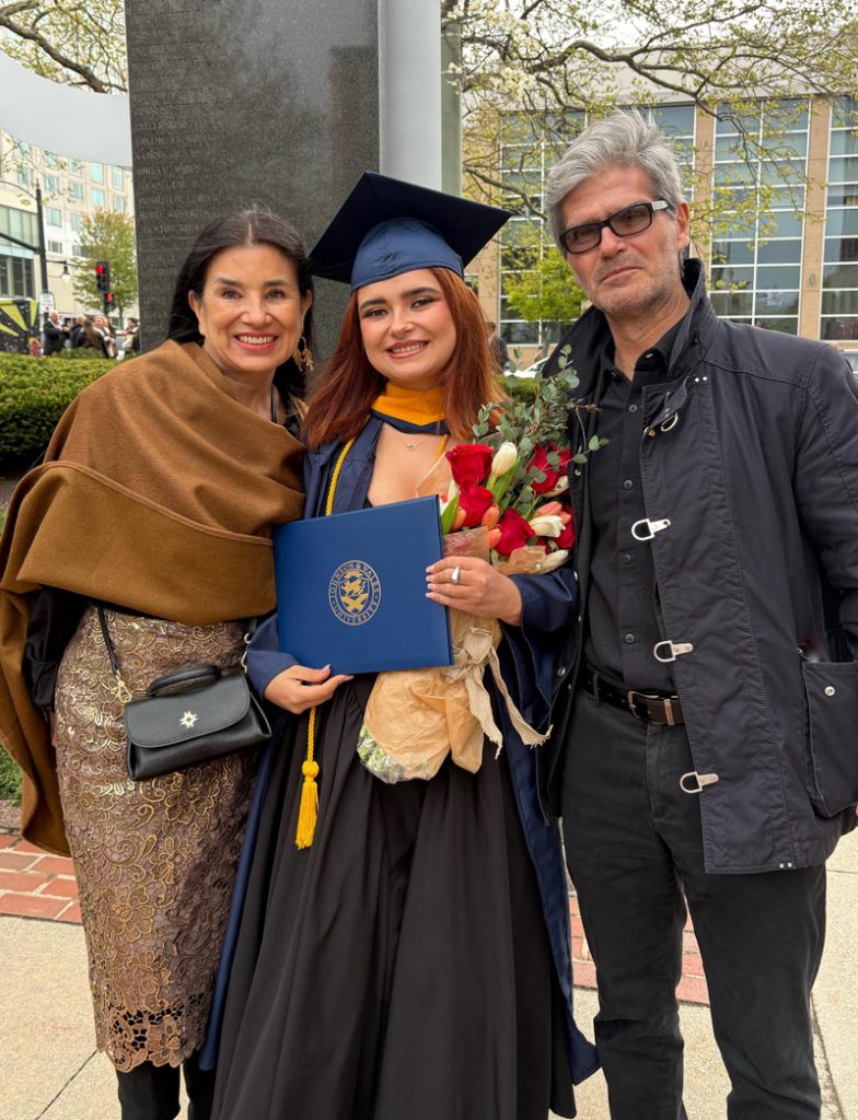 Hannah Levy in her cap and gown, holding her diploma and flowers, with her mother and father outside after the commencement ceremony at the Amica Mutual Pavilion.