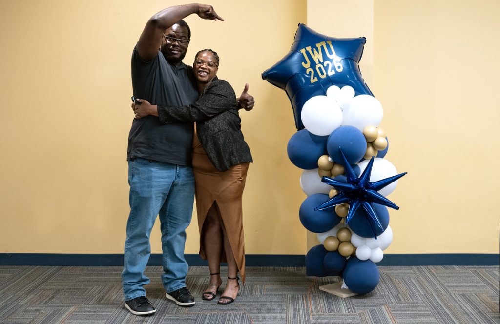 Jennifer Butler hugs her husband, who points to the balloon decoration embossed with “JWU 2026” in gold lettering, at the JWU Online Senior Toast held in the Wildcat Center on the afternoon before commencement.