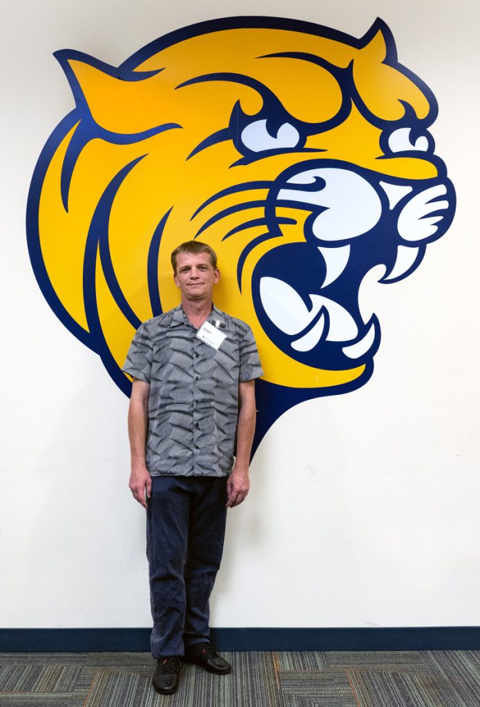 Joshua Orndorff standing in front of a painting of a wildcat on the wall at the JWU Online Senior Toast held in the Wildcat Center on the afternoon before commencement.