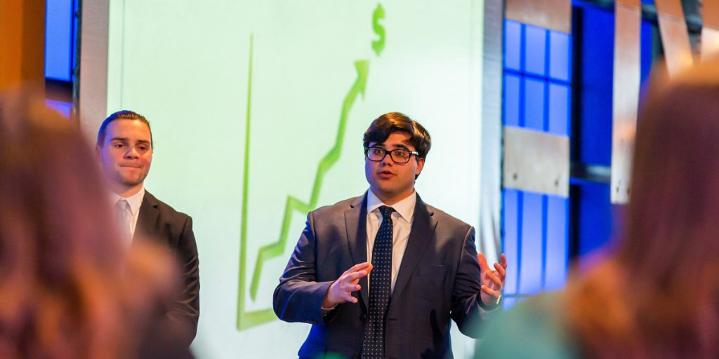 Young man wearing glasses, suit and a tie, standing in front of a board displaying an ascending arrow pointing to a dollar sign, speaking to a group of people.