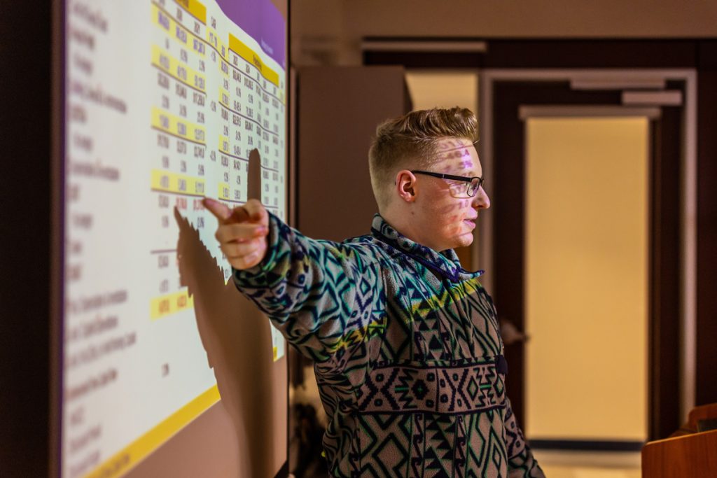Male with glasses seen in sideview pointing at numerical information projected on a board behind him.