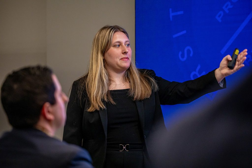 Woman standing in front of an audience holding a remote clicker for the slide presentation displayed behind her.