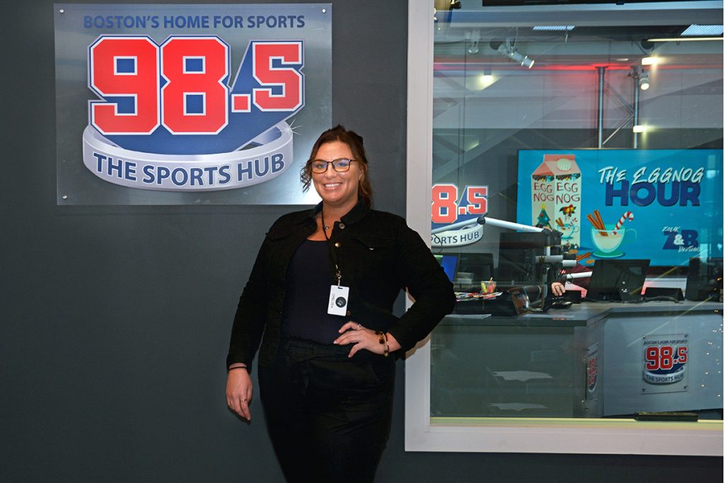 Chelsea Delfino, a female student in the SEEM program, stands with hand on hip alongside a logo on the wall for 98.5 The Sports Hub and the window that looks into the broadcast studio.
