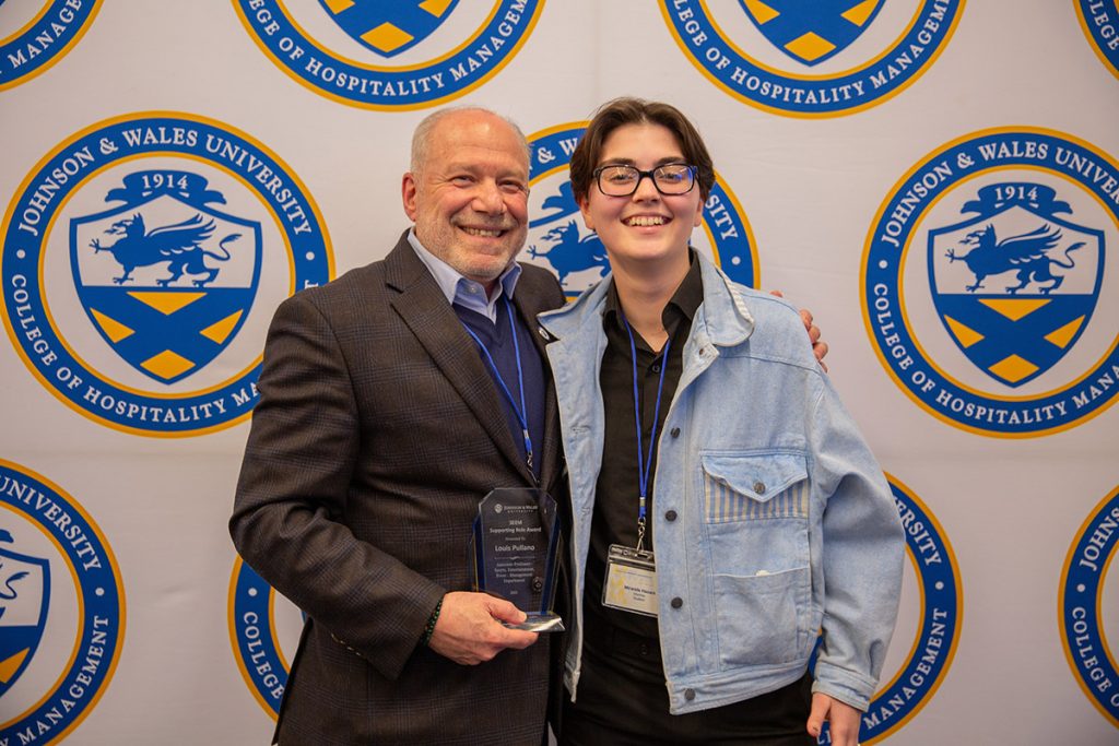 Professor Louis Pullano with a Johnson & Wales student photographed against a backdrop of JWU College of Hospitality logos.