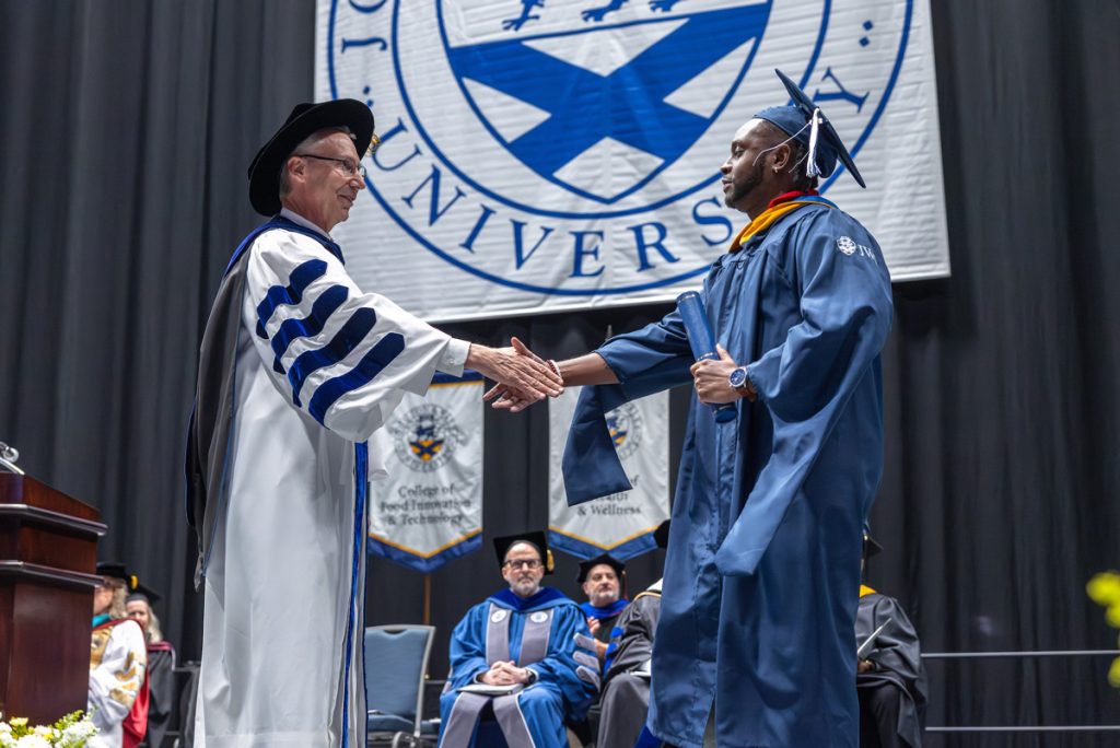 President Mathieu shaking the hand of a male graduate as he walks across the stage at the Charlotte Commencement.