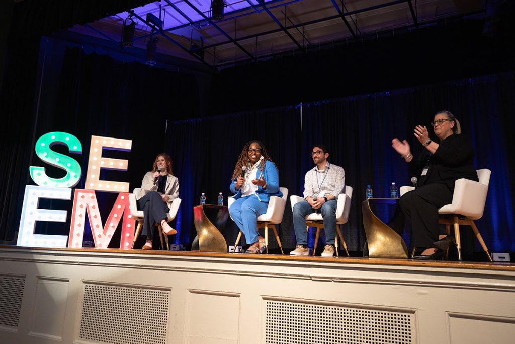 Two females and one male holding microphones are seated on a stage with a a woman who is moderating the SEEM event.