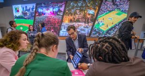 Gentleman in sports jacket showing a group of students his laptop screen against the backdrop of large, colorful photos of JWU sports events on the SEEM Lab wall.