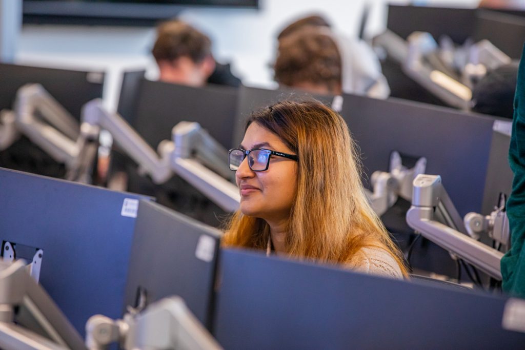 Young smiling woman peering over the row of computers in a classroom, with a group of male students at computers in the background.