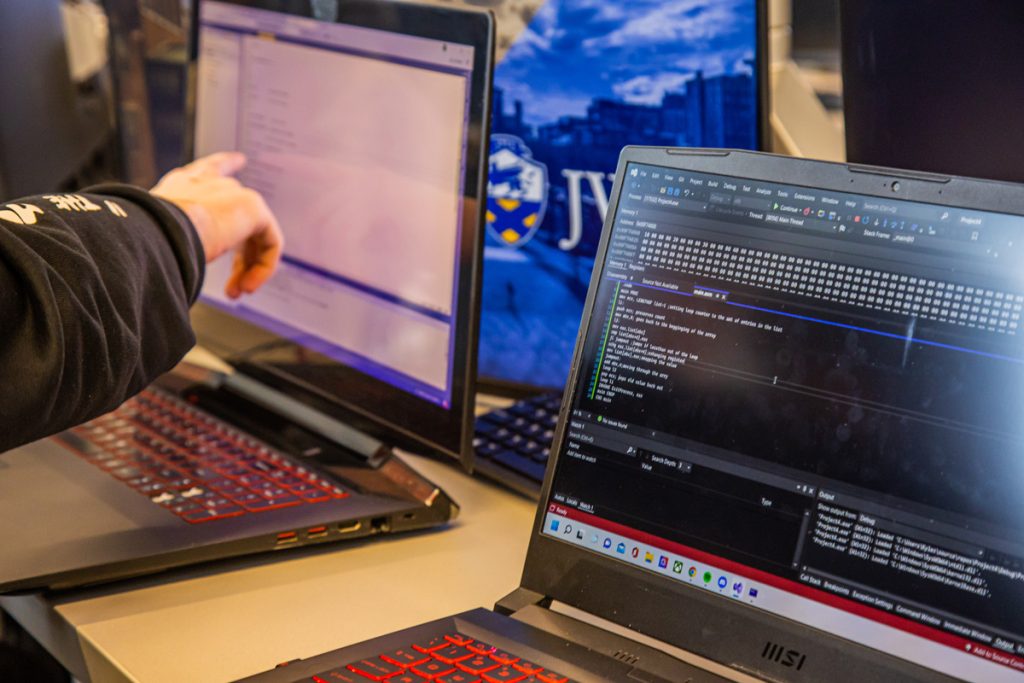 View of two side-by-side laptop screens, with someone's hand pointing at one, while a JWU logo is visible on a screen in the background.