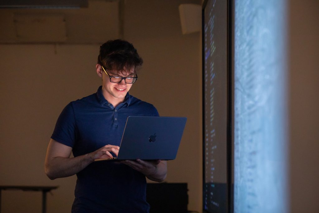 Young smiling man with glasses standing next to a projection screen in a classroom while looking at his laptop screen.