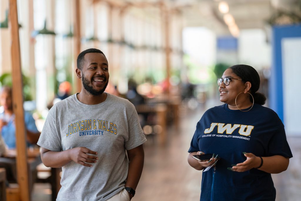 Young man and woman of color, wearing JWU t-shirts, smiling and talking as they walk side-by-side through a hallway.