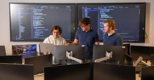 Three male students standing in a computer lab looking at a laptop screen with two large screens on which data is projected behind them.