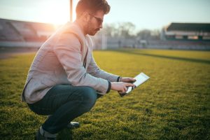Man in a blazer crouching on a sports field at sunset, checking data on a tablet beside the field markings