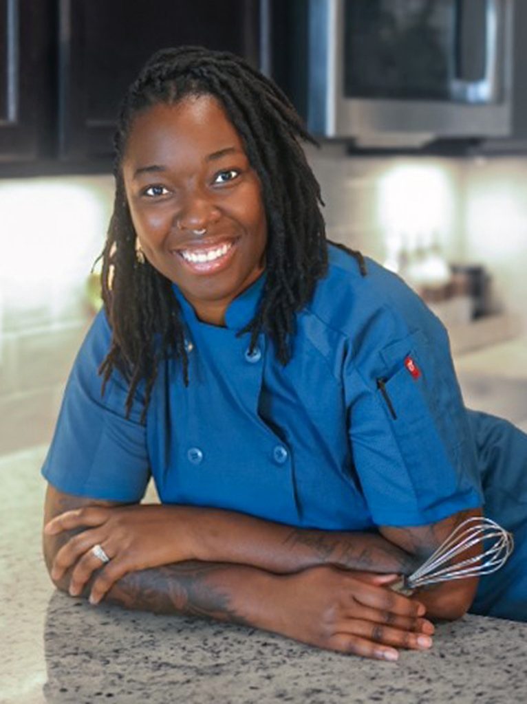 Portrait of Direka Gordon, smiling while holding a whisk and leaning on the countertop in her kitchen.