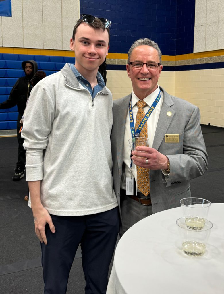 Tyler Power in casual clothing and sunglasses on his head stands with JWU President, Joe Greene, during the Senior Toast at the JWU Wildcat Center.