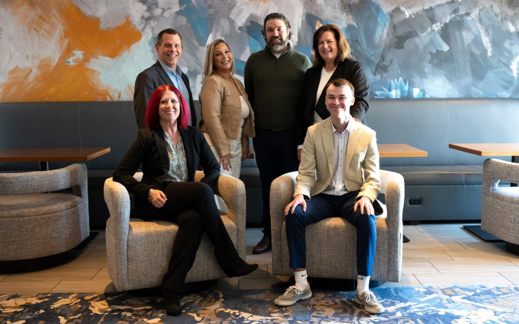 Power and five members of his mentorship team (two males and three females) are seated in a lounge area with a blue and orange abstract mural on the wall behind them, at the Marriott Quincy.