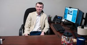 Young man, Tyler Power, seated in his new office, with computer screen, phone and candy jar on the desk, at the Marriott Quincy