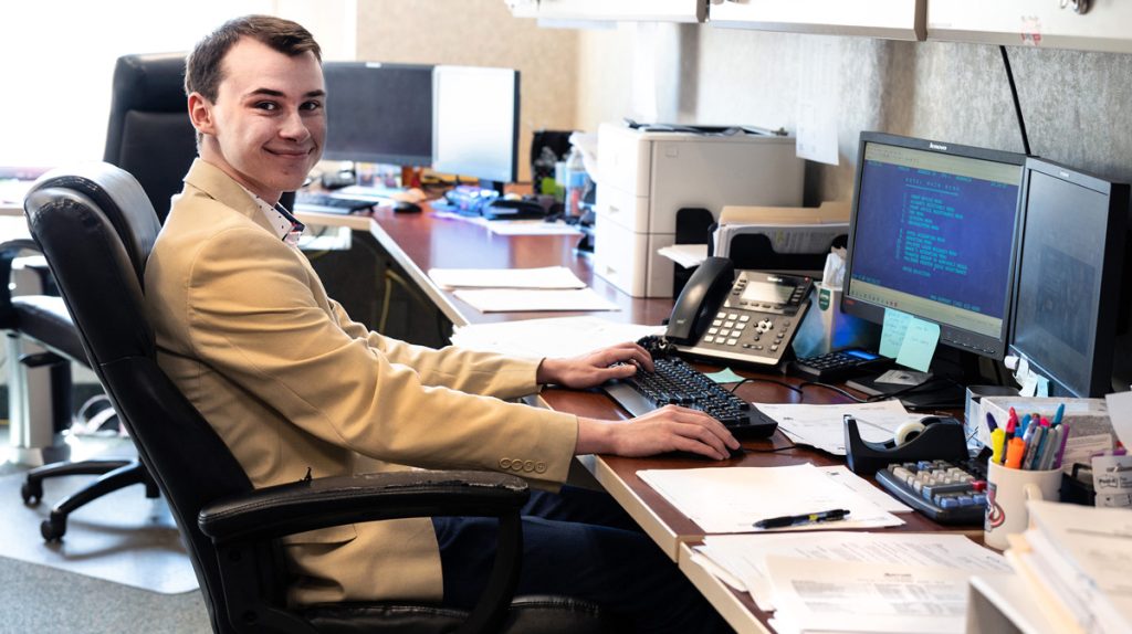 Power seated at a desk, with a phone and two computer monitors and additional monitors in the space behind him.