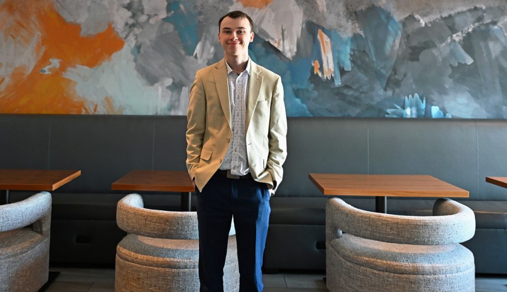 Tyler Power wearing a tan casual business jacket and dark pants, standing in front of a mural, and upholstered chairs and tables in the concierge lounge at the Marriott Boston Quincy Hotel. 