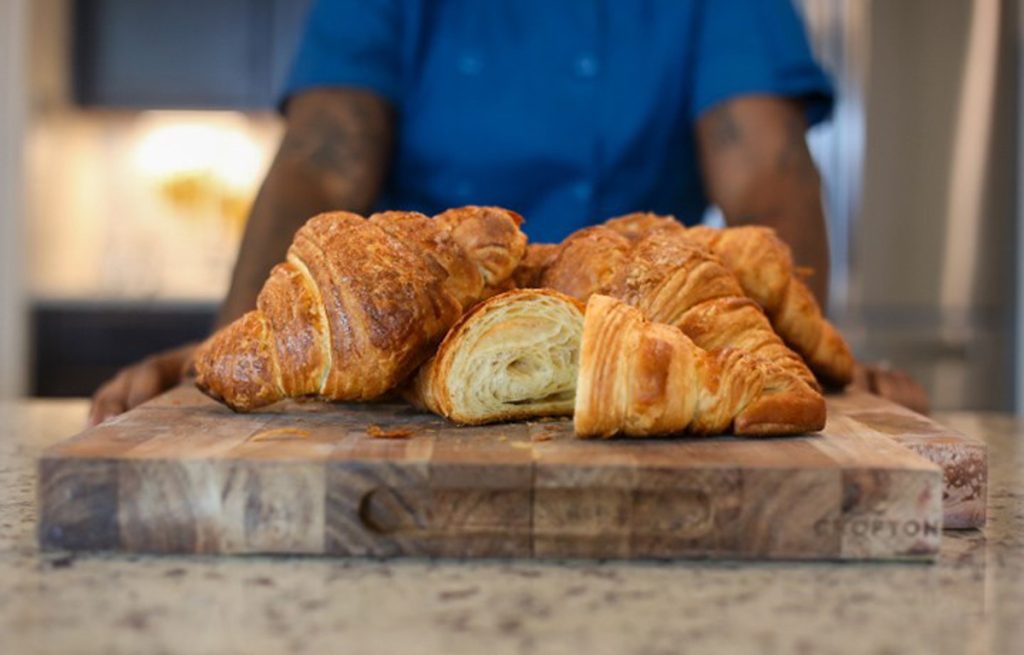 Freshly baked croissants on a wooden cutting board with the torso and arms of Direka Gordon in her blue uniform in the background.