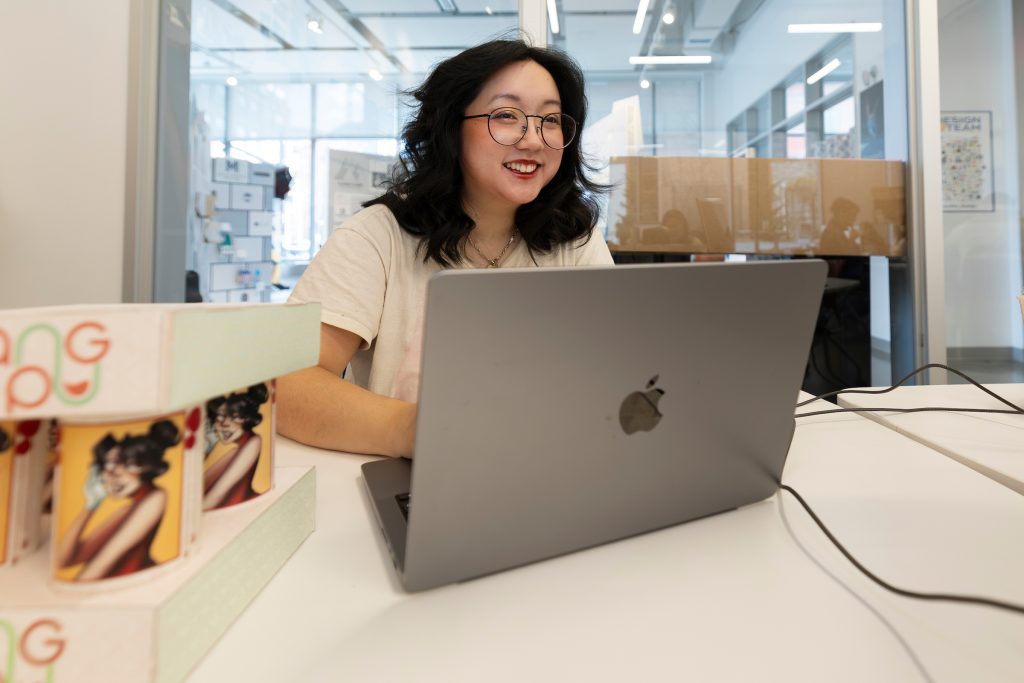 A female student wearing glasses and smiling is seated at a table in a university building with a laptop open in front of her,