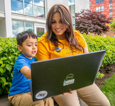 Emmeline Orellana and her son, Jeremy, are seated outdoors. viewing a large laptop screen that Emmeline is holding..