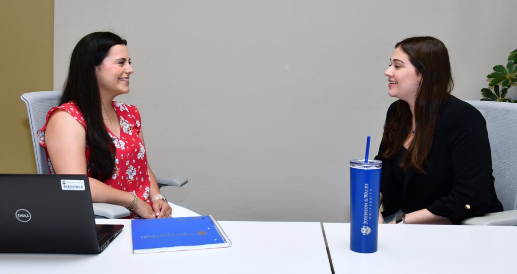 Two women, one with a laptop and notebook, are smiling as they sit facing one another at a table.