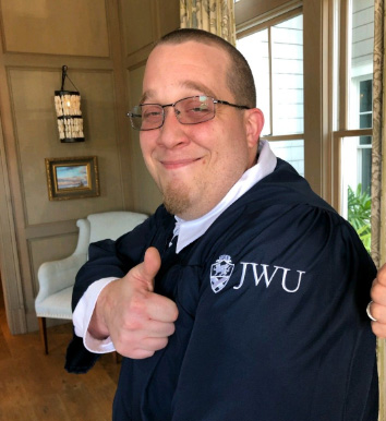 Michael Yates in commencement gown with the JWU logo visible, is giving "thumbs up."