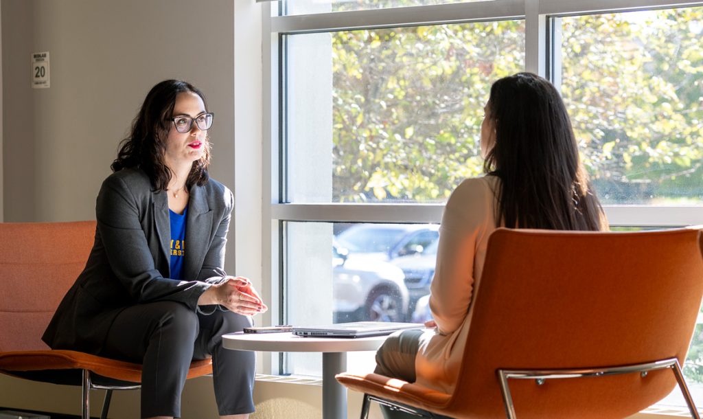 Seated in front of a window, a female staff member listens to a student. who is facing her