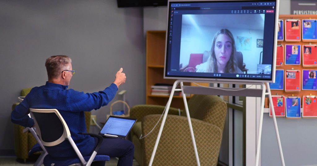 Professor Jeff Binczyk is seated and giving "thumbs up" to a young woman on a screen during a virtual meeting.