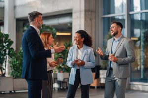 Four business professionals in an office lobby, smiling and laughing while talking together, two holding coffee cups.