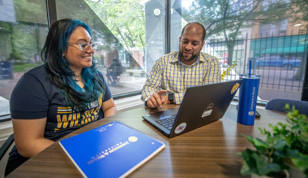 A female student and a male staff member are seated at a table in front of windows, and are looking at the staff member's laptop screen.